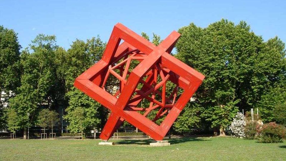 Grande scultura astratta in metallo rosso, composta da telai cubici inclinati e intrecciati, collocata su un prato. Sullo sfondo si vede un’area verde con alberi ad alto fusto e cielo sereno.
