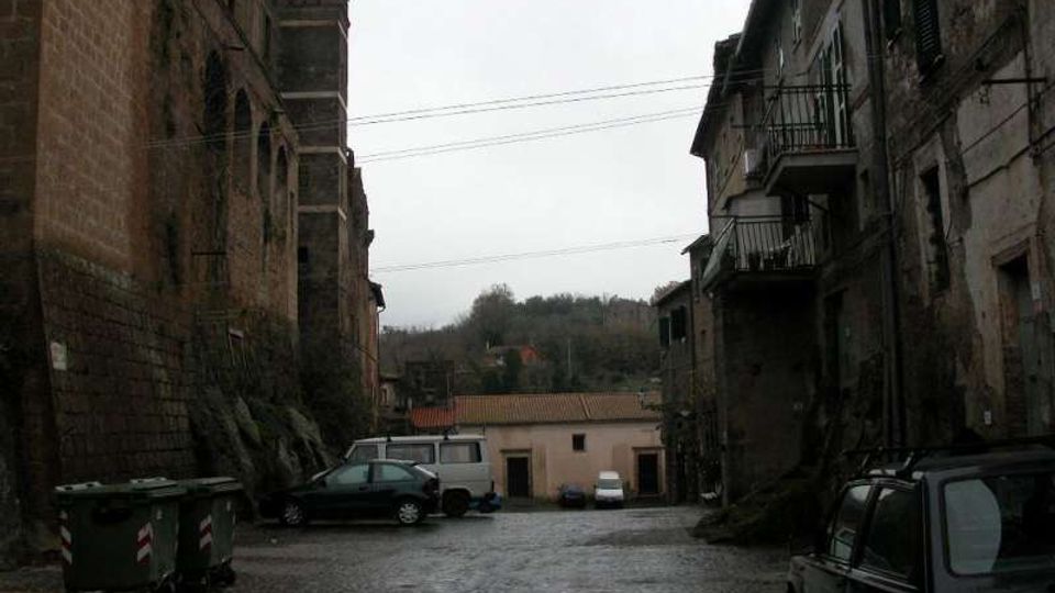 Piazzale lastricato in pietra tra edifici in muratura antica, con facciate scure, finestre e balconi in ferro. Sullo sfondo si vede un edificio basso con tetto in tegole e una collina alberata, con auto parcheggiate e cassonetti lungo i lati.
