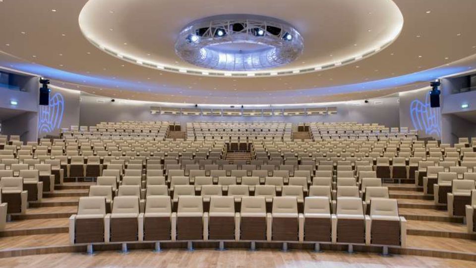 Ampio auditorium moderno con file di poltrone beige disposte a gradoni e pavimento in legno. Il soffitto curvo presenta un grande elemento circolare con illuminazione integrata e pannelli laterali con proiezioni luminose.
