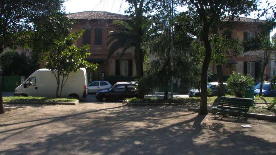 Piccolo parco urbano con alberi alti che proiettano ombre su un’area in terra battuta e una panchina verde. Sullo sfondo si vedono auto parcheggiate e un edificio in mattoni a due piani con tetto in tegole, parzialmente nascosto dalla vegetazione.
