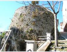 Grande struttura circolare in pietra grezza, simile a una torre o bastione fortificato, vista dal basso. In primo piano ci sono scale e muretti in cemento con ringhiere, un albero spoglio e un’area in pendenza accanto a edifici.