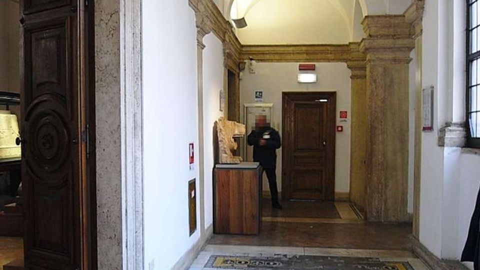 Corridoio interno con soffitto a volte e archi in muratura, pareti bianche e pavimento in mosaico decorato. Ai lati si vedono porte in legno scuro e una grande finestra, con un banco espositivo vicino al fondo.