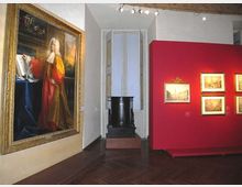 Interno di una sala museale con pavimento in parquet scuro e illuminazione a binario: a sinistra un grande dipinto incorniciato, a destra una parete espositiva rossa con quadri più piccoli. Sul fondo si vede una nicchia con una finestra alta e un basamento scuro.