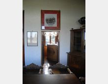 Interno di una sala con tavolo e sedie in legno intagliato, una credenza vetrata sulla destra e quadri appesi a una parete chiara. Al centro, una porta aperta conduce a un’area illuminata con finestra e tende, con vista sul verde esterno.