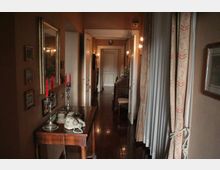 Lungo corridoio interno con pavimento in legno lucido e pareti color beige, illuminato da applique e con porte bianche lungo i lati. In primo piano un tavolino in legno con specchio, candelabri e telefono, mentre sul lato destro ci sono tende pesanti davanti alle finestre.