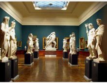 Sala di museo con pareti verde scuro e lucernario centrale, pavimento in legno lucido e sculture in marmo su piedistalli disposte lungo il percorso. Dipinti incorniciati alle pareti e una grande scultura al centro della sala.