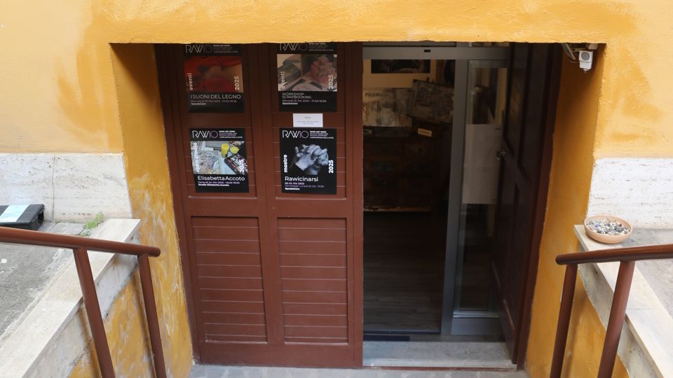 Scala esterna in pietra che scende a un ingresso seminterrato incassato in una parete gialla, con corrimano metallici ai lati. In fondo si vede una porta marrone con locandine e una porta vetrata scorrevole.
