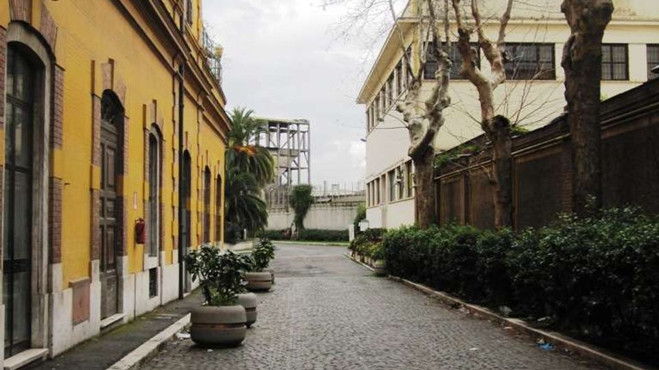 Strada acciottolata in un cortile urbano, con un lungo edificio giallo con persiane e portoni sulla sinistra e un edificio chiaro sulla destra. Lungo il percorso ci sono grandi vasi con piante, una fila di alberi spogli e siepi vicino a un muro, sotto un cielo nuvoloso.