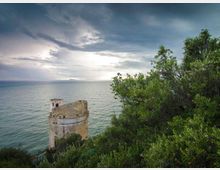 Torre costiera in pietra su un promontorio roccioso, circondata da vegetazione mediterranea, con vista sul mare. Sullo sfondo si vede l’orizzonte marino sotto un cielo nuvoloso e luminoso.