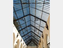 Interno di una galleria coperta con tetto a falde in vetro sorretto da una struttura metallica scura con elementi decorativi. Lungo i lati si vedono pareti chiare con archi e grandi finestre, illuminate dalla luce naturale che filtra dal lucernario.