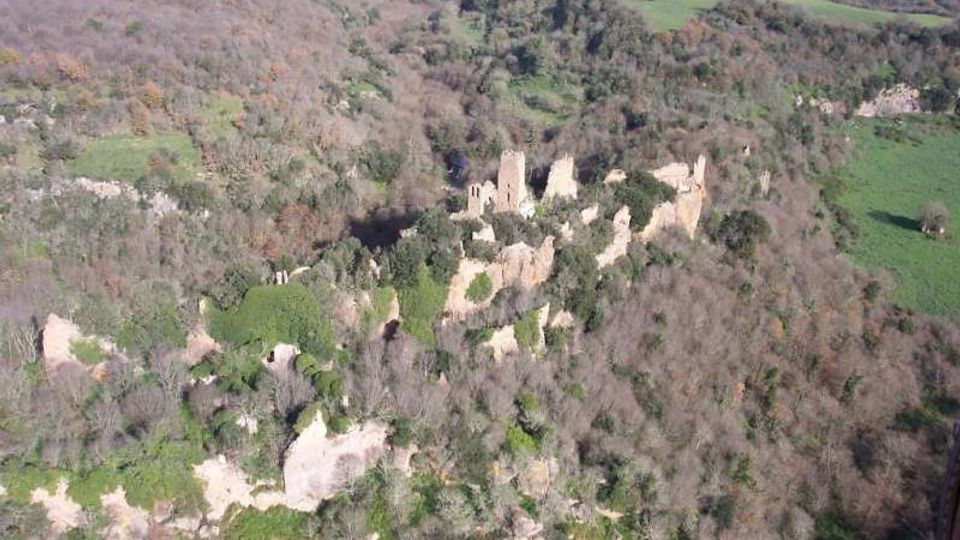 Veduta aerea di un complesso di rovine in pietra, con resti di torri e mura, immerso in una fitta area boschiva. Intorno si vedono pendii rocciosi e campi agricoli che si estendono nella campagna circostante.