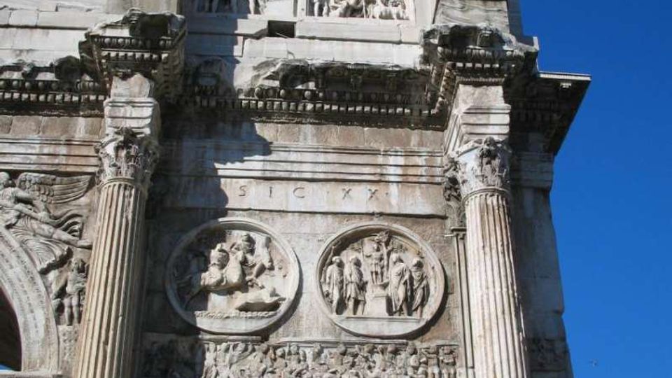 Vista ravvicinata di un arco monumentale romano in pietra, riccamente decorato con rilievi e iscrizioni, con due grandi colonne scanalate ai lati e nicchie con statue nella parte superiore. Sullo sfondo si vede un cielo azzurro limpido.
