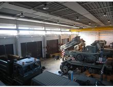 Interno di un grande capannone industriale adibito a officina, con soffitto in metallo e ampie finestre alte lungo le pareti. Nella zona centrale sono presenti mezzi pesanti e macchinari in manutenzione, con attrezzature e materiali disposti sul pavimento.