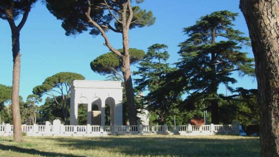Parco erboso con alti pini e cedri, con un piccolo edificio bianco con archi e una balaustra in pietra sullo sfondo, sotto un cielo sereno.