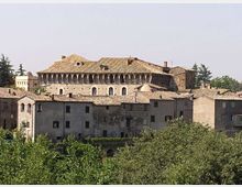 Veduta panoramica di un centro storico con edifici in pietra e tetti in coppi, dominato da un grande palazzo antico con loggiato ad archi e copertura a falde. In primo piano si vedono alberi e vegetazione che incorniciano le case addossate tra loro.