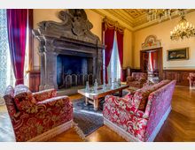 Salone storico in stile classico con grande camino monumentale in pietra scolpita, soffitto decorato e lampadari. Arredi in tessuto rosso a motivi floreali attorno a un tavolino, pavimento in parquet, alte finestre con tende rosse e boiserie in legno lungo le pareti.