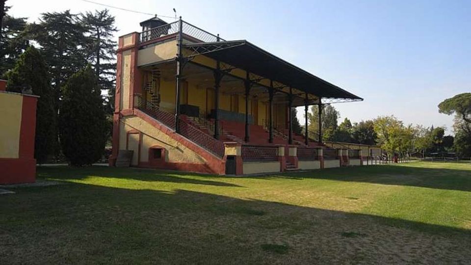 Tribuna in muratura e mattoni con gradinate e ringhiere, coperta da un’ampia tettoia metallica sostenuta da pilastri. La struttura si affaccia su un campo erboso ed è circondata da alberi in un’area verde.