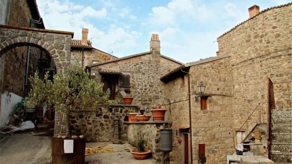 Cortile in un borgo con edifici in pietra e tetti in coppi, con scale esterne, piccoli balconi e lampioni a muro. Al centro ci sono vasi in terracotta e una pianta in una fioriera, su pavimentazione in pietra sotto un cielo parzialmente nuvoloso.