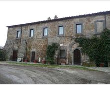 Facciata di un casale rurale in pietra a due piani, con tetto in coppi e finestre rettangolari incorniciate. Al piano terra ci sono porte in legno, un portale ad arco e piante rampicanti con grandi vasi lungo la base, affacciati su una strada sterrata.