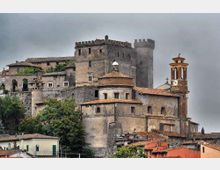 Castello medievale in pietra su un’altura, con mura merlate, una torre cilindrica e vari edifici annessi dai tetti in tegole. In primo piano si vedono case del borgo e, sul lato destro, un campanile con cuspide.