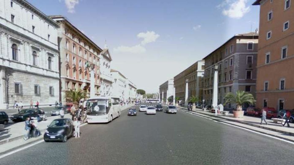 Ampio viale cittadino con carreggiata a più corsie e traffico, fiancheggiato da edifici storici in muratura di diversi piani. Marciapiedi larghi con file di auto in sosta, palme in grandi fioriere e cielo sereno sullo sfondo.
