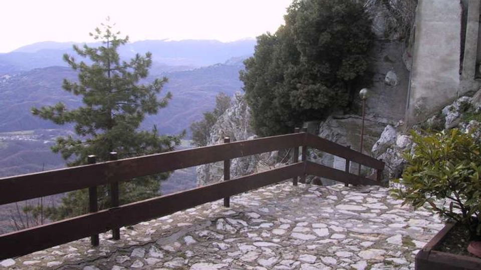 Terrazza panoramica con pavimentazione in pietra irregolare e parapetto in legno, affacciata su una valle montuosa. A lato si vedono parete rocciosa, alberi e un edificio in muratura addossato alla scogliera.