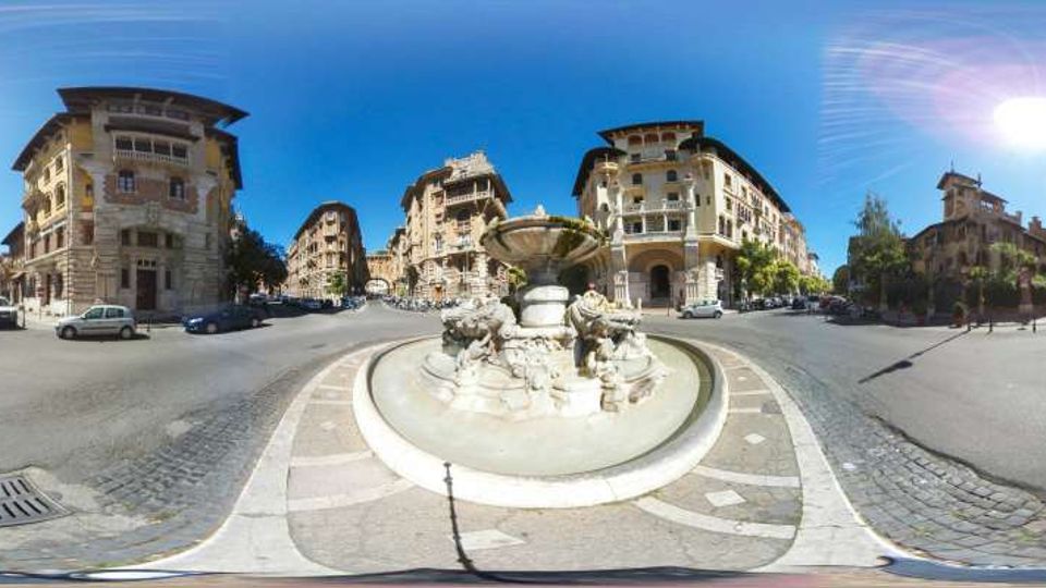 Grande piazza urbana con una fontana monumentale in pietra al centro di una rotonda, circondata da ampie strade asfaltate. Intorno si vedono palazzi storici di più piani con facciate decorate e balconi, sotto un cielo sereno e luminoso.