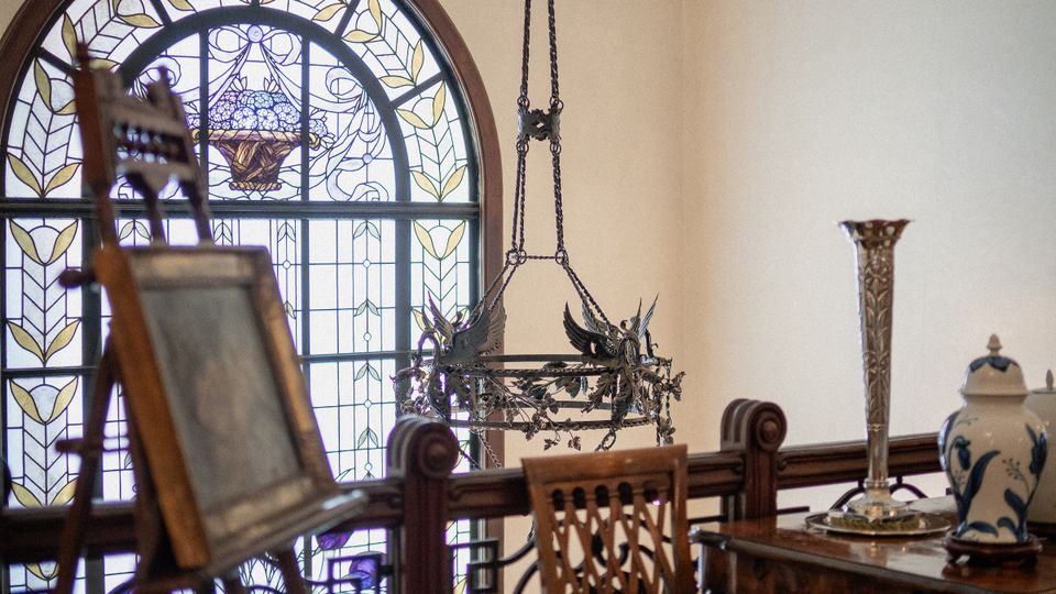 Soppalco interno in stile d’epoca con grande finestra ad arco in vetro colorato e ringhiera in ferro battuto decorata. Nello spazio si vedono un lampadario sospeso, un cavalletto in legno e un piccolo tavolo con sedia e oggetti decorativi.