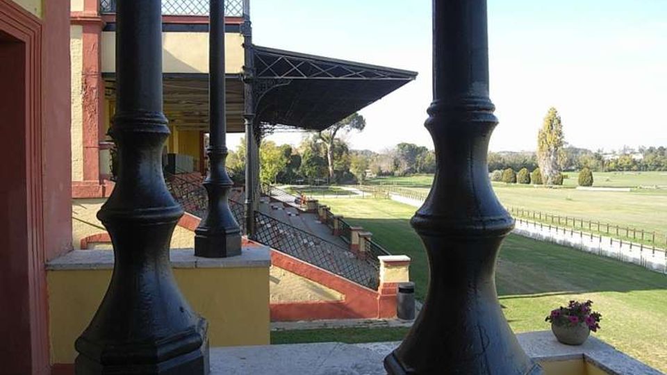 Vista da una tribuna coperta con colonne in ghisa e struttura metallica, affacciata su una pista erbosa recintata e ampi campi verdi. A sinistra si vede l’edificio della tribuna con pareti color ocra e rosso e gradinate sottostanti.