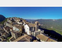 Veduta aerea di un borgo collinare con case in pietra dai tetti in tegole addossate tra loro e una chiesa con campanile al centro. Sullo sfondo si vedono vallate e montagne verdi sotto un cielo limpido.