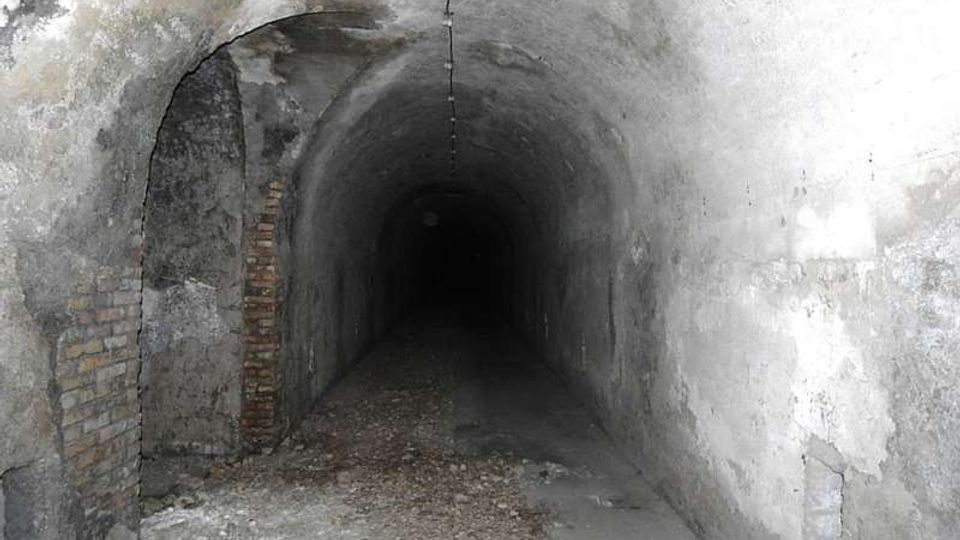 Tunnel sotterraneo stretto con volta a botte in cemento grezzo, pareti macchiate e tratti di mattoni a vista sul lato sinistro. Il pavimento è parzialmente coperto da detriti e il corridoio si estende in profondità verso un’area più buia.