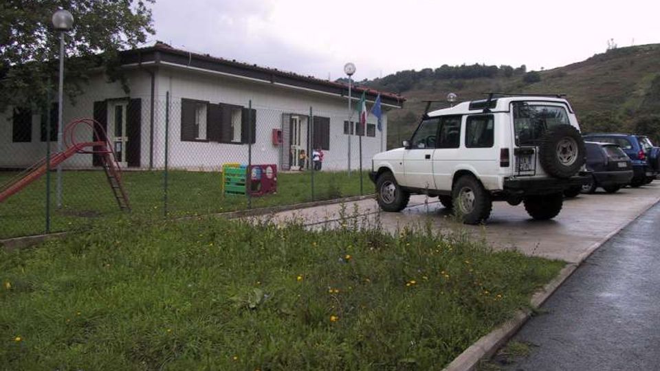 Edificio basso a un piano con facciata chiara e persiane scure, circondato da una recinzione metallica e da un’area verde con piccolo parco giochi. In primo piano si vede un parcheggio con auto, sullo sfondo colline e cielo nuvoloso.