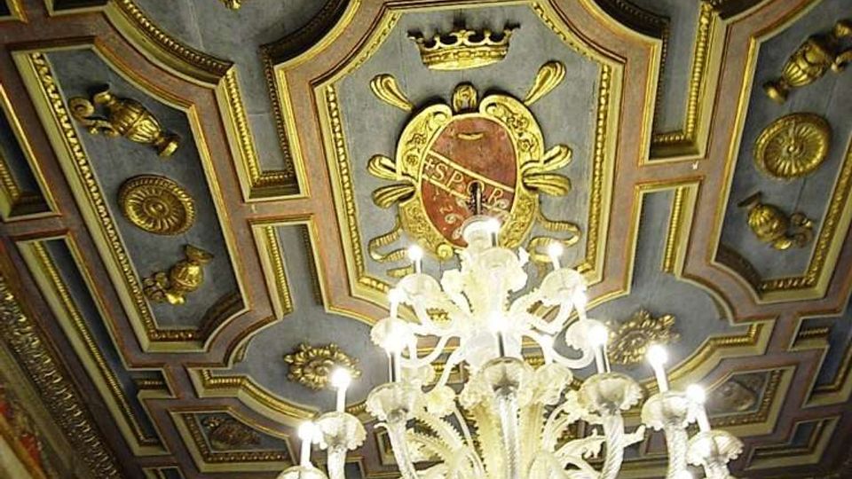 Soffitto a cassettoni decorato con cornici e rosoni dorati, con uno stemma centrale, in un ambiente interno di stile storico. Al centro è sospeso un grande lampadario in vetro con molte braccia illuminate, sopra pareti affrescate e finestre.