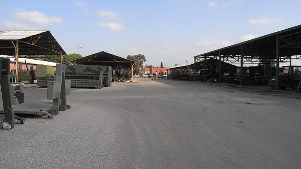 Ampio piazzale asfaltato in un’area industriale, con grandi tettoie metalliche aperte ai lati che coprono attrezzature e veicoli. Sullo sfondo si vedono altri capannoni e un cielo parzialmente nuvoloso.