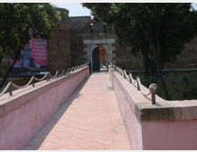 Passerella pedonale in mattoni con parapetti intonacati e catene, che conduce a un portale ad arco in pietra su un muro di fortificazione in mattoni. Ai lati si vedono alberi e un grande cartellone, con vegetazione che circonda l’ingresso.