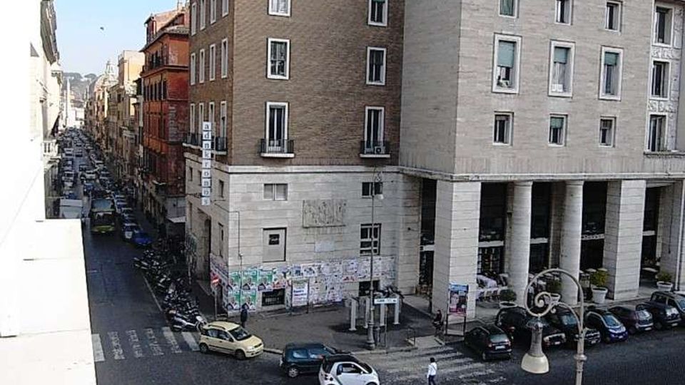 Incrocio urbano con strisce pedonali e traffico, visto dall’alto, affiancato da edifici in muratura e pietra. In primo piano un grande palazzo con portico a colonne e facciate con finestre rettangolari lungo una strada stretta fiancheggiata da palazzi.