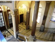 Interno di un atrio storico con scale in pietra e pavimento a motivi geometrici in marmo, illuminato da grandi finestre. Colonne massicce e archi decorati incorniciano porte in legno e un ingresso con cornice ornamentale.