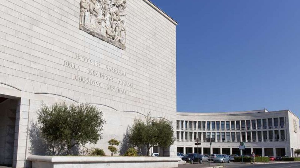 Complesso di edificio istituzionale in pietra chiara con grande facciata liscia, bassorilievo e iscrizione incisa; sullo sfondo un’ala curva con file di finestre. Davanti si vedono un’aiuola con alberi, un parcheggio con auto e una strada sotto cielo sereno.