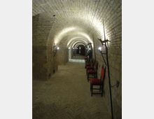 Corridoio sotterraneo in pietra con volta a botte in mattoni, illuminato da lampade a parete lungo entrambi i lati. Sul lato destro sono allineate sedie con seduta rossa e sono appoggiate lunghe armi ad asta contro il muro, mentre il passaggio prosegue fino a una porta in fondo.