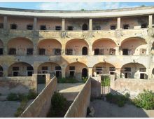 Cortile interno di un edificio storico in rovina, con struttura semicircolare a più piani di arcate e aperture murate o sbarrate. In primo piano si vedono muretti bassi che delimitano corridoi e aree con vegetazione spontanea.