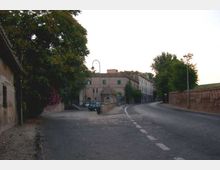 Strada asfaltata che curva a destra accanto a un marciapiede in pietra, con muri di mattoni e vegetazione ai lati. Sullo sfondo si vedono edifici in muratura chiara, un lampione ad arco e alcuni veicoli parcheggiati vicino a un piccolo accesso.