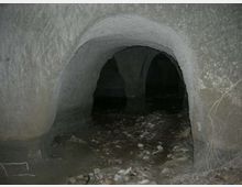 Ingresso ad arco di una grotta o galleria sotterranea con pareti in roccia grezza. Il pavimento è irregolare, coperto di sassi e detriti, e l’interno si apre in più passaggi bui.