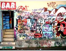 Ingresso di un bar con insegna “BAR” e gradini che scendono verso una porta aperta, con scaffali di bottiglie visibili all’interno. Accanto, un grande muro esterno ricoperto di graffiti multicolori, scritte e poster pubblicitari sovrapposti.