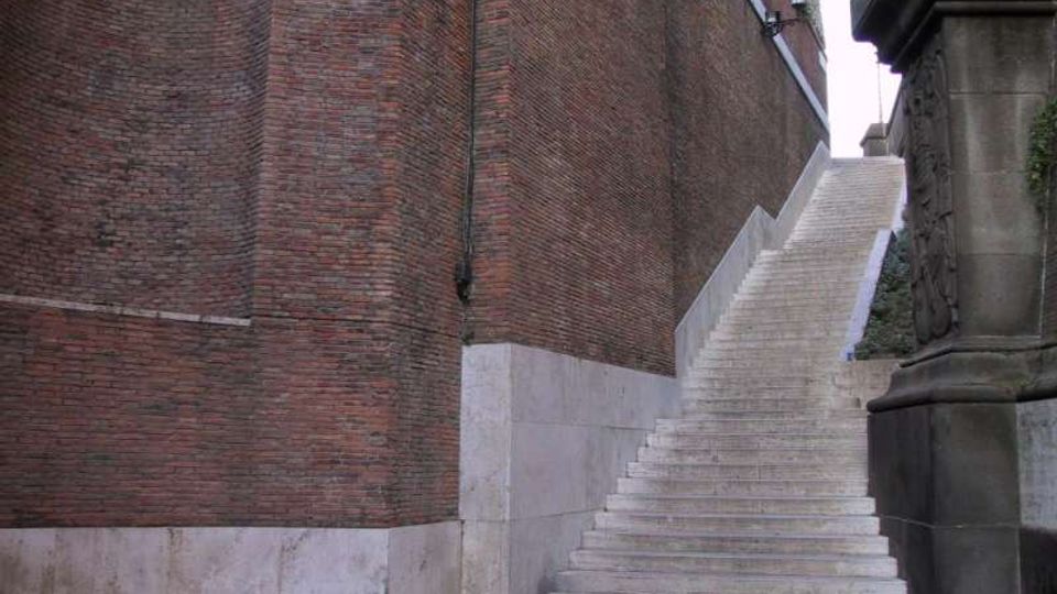 Scalinata in pietra chiara che sale lungo alte mura di mattoni rossi, con basamento in pietra e un canale di scolo verticale sulla parete. A destra si vede un grande piedistallo in pietra scolpita che costeggia i gradini, in un passaggio urbano stretto.