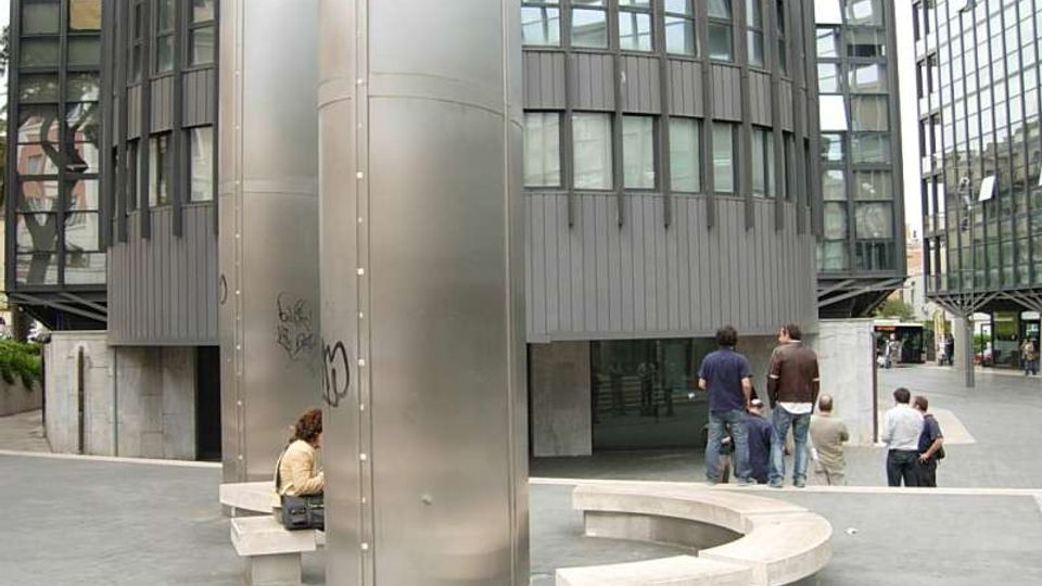 Piazza urbana moderna con due edifici cilindrici in vetro e metallo e una grande colonna centrale rivestita in acciaio. Nell’area pedonale ci sono panchine curve in cemento e facciate vetrate che circondano lo spazio.