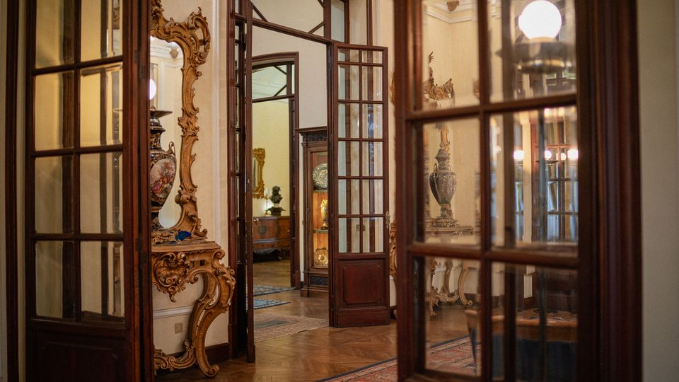 Interno di un palazzo storico con porte a vetri e telai in legno scuro che si aprono su stanze comunicanti. Si vedono pavimento in parquet, tappeti e arredi d’epoca con specchi e consolle intagliate e vasi decorativi.