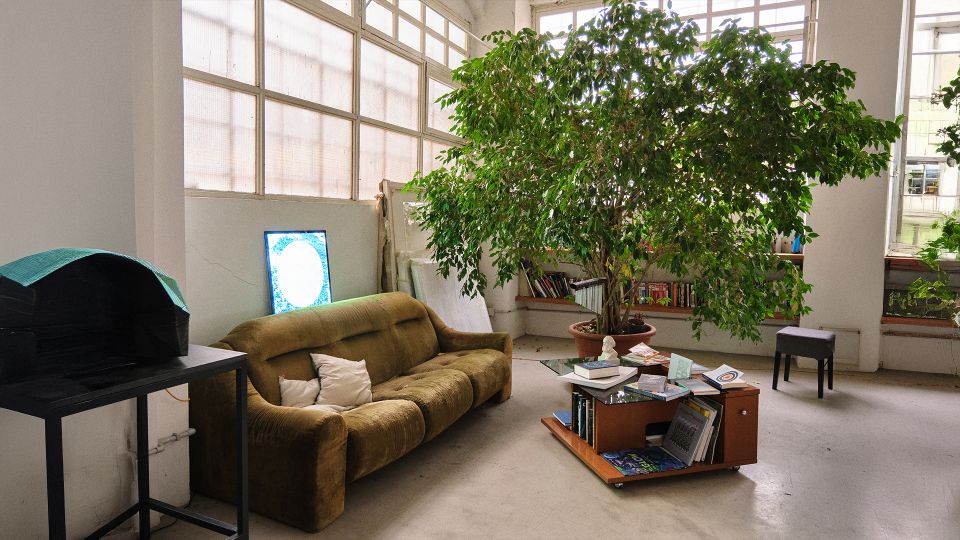 Ampio interno in stile loft con grandi finestre a riquadri che illuminano un’area relax con divano e tavolino basso pieno di libri. Una grande pianta in vaso domina la stanza, con scaffali e arredi sparsi su pavimento in cemento.