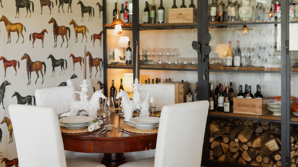 Sala da pranzo con tavolo rotondo in legno apparecchiato e sedie bianche, decorata con candelabri e centrotavola a forma di cavallo. Sullo sfondo, grande armadio vetrato con bottiglie di vino, bicchieri e legnaia in basso, e parete con carta da parati raffigurante cavalli.