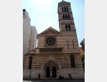 Facciata di una chiesa in stile romanico-gotico con pietra a fasce orizzontali, grande rosone centrale e portale ad arco ogivale. Accanto si innalza un campanile alto con logge ad archi sovrapposti, in un contesto urbano tra edifici moderni.