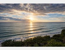 Vista panoramica dell’oceano al tramonto, con onde regolari che arrivano alla riva e riflesso dorato del sole sull’acqua sotto un cielo parzialmente nuvoloso. In primo piano si vedono vegetazione costiera e una staccionata con lampione lungo un sentiero sopraelevato.